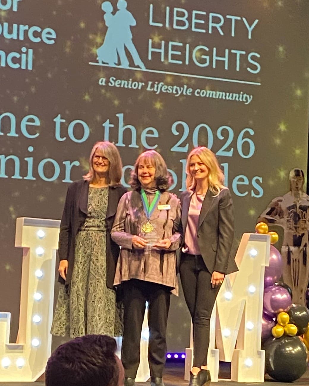 Three women on stage at the 2026 Joe Henjum Senior Accolades. The woman in the center wears a medal and holds the award.