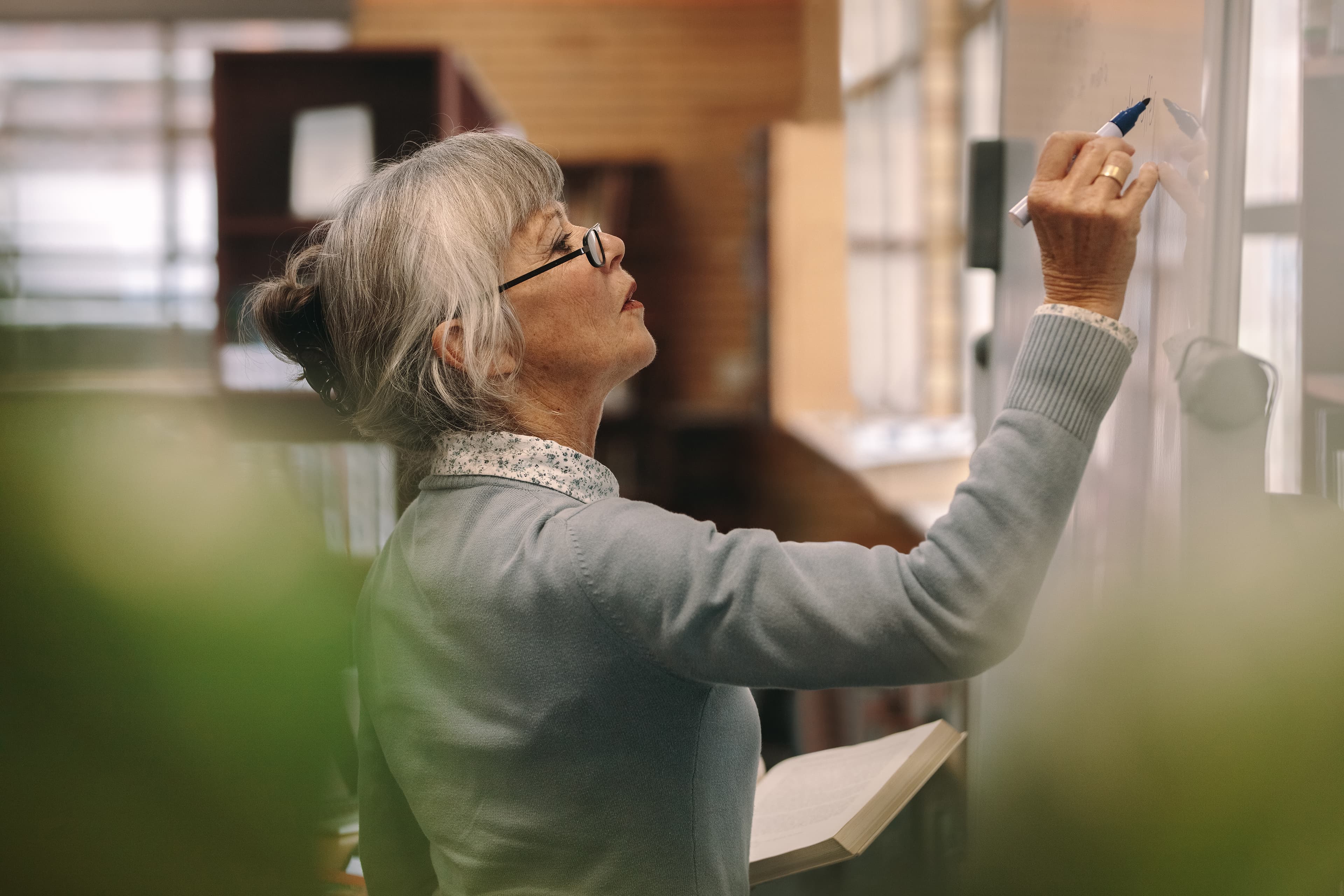 An older woman with gray hair and glasses writes on a whiteboard, holding an open book