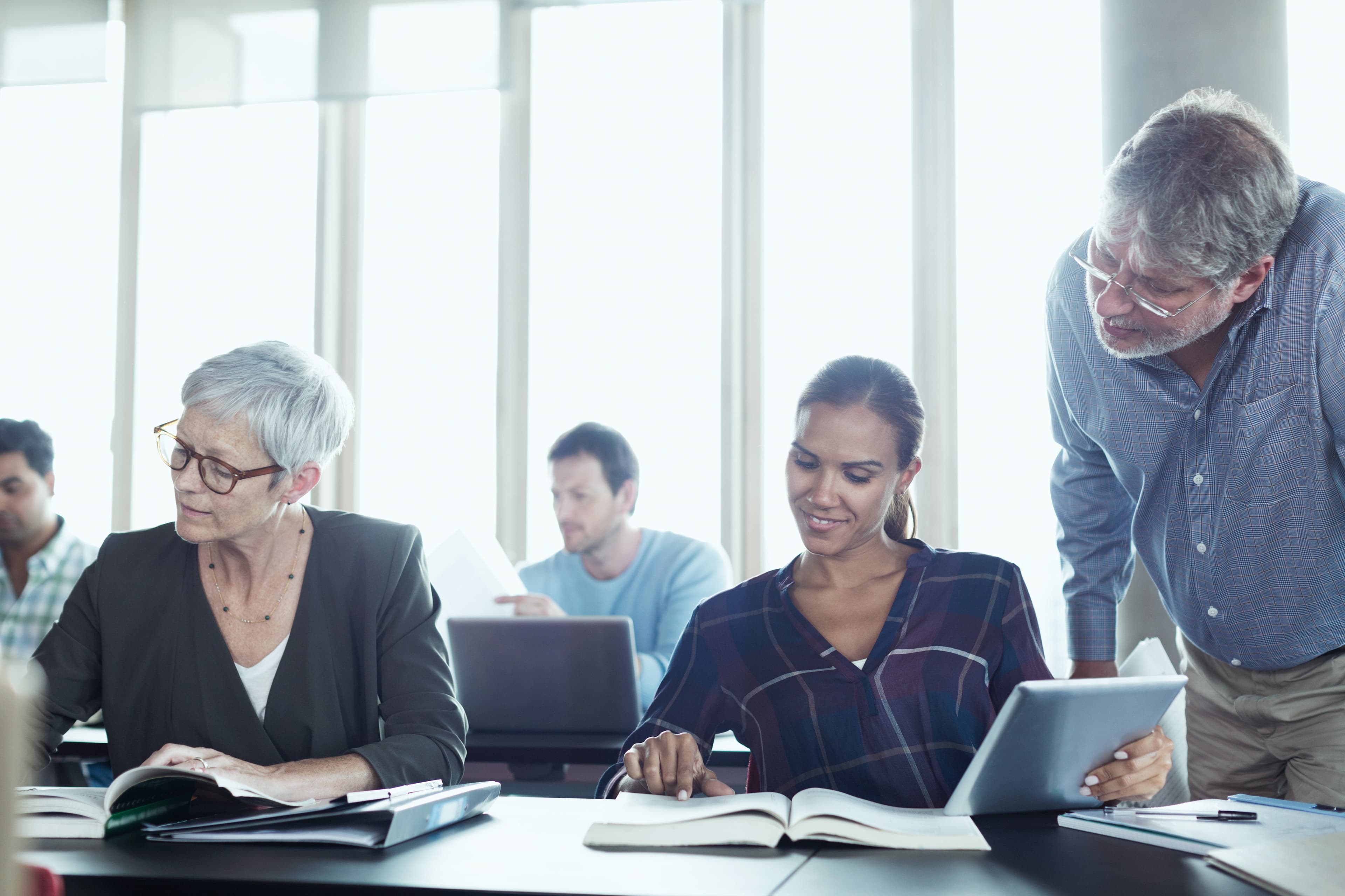An instructor with gray hair leans in to help two adult learners reading at their desks in a bright classroom